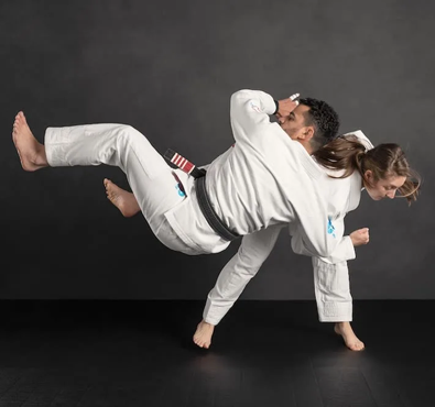 Two women practicing a Brazilian Jiu Jitsu throw technique in white gis against a dark studio background