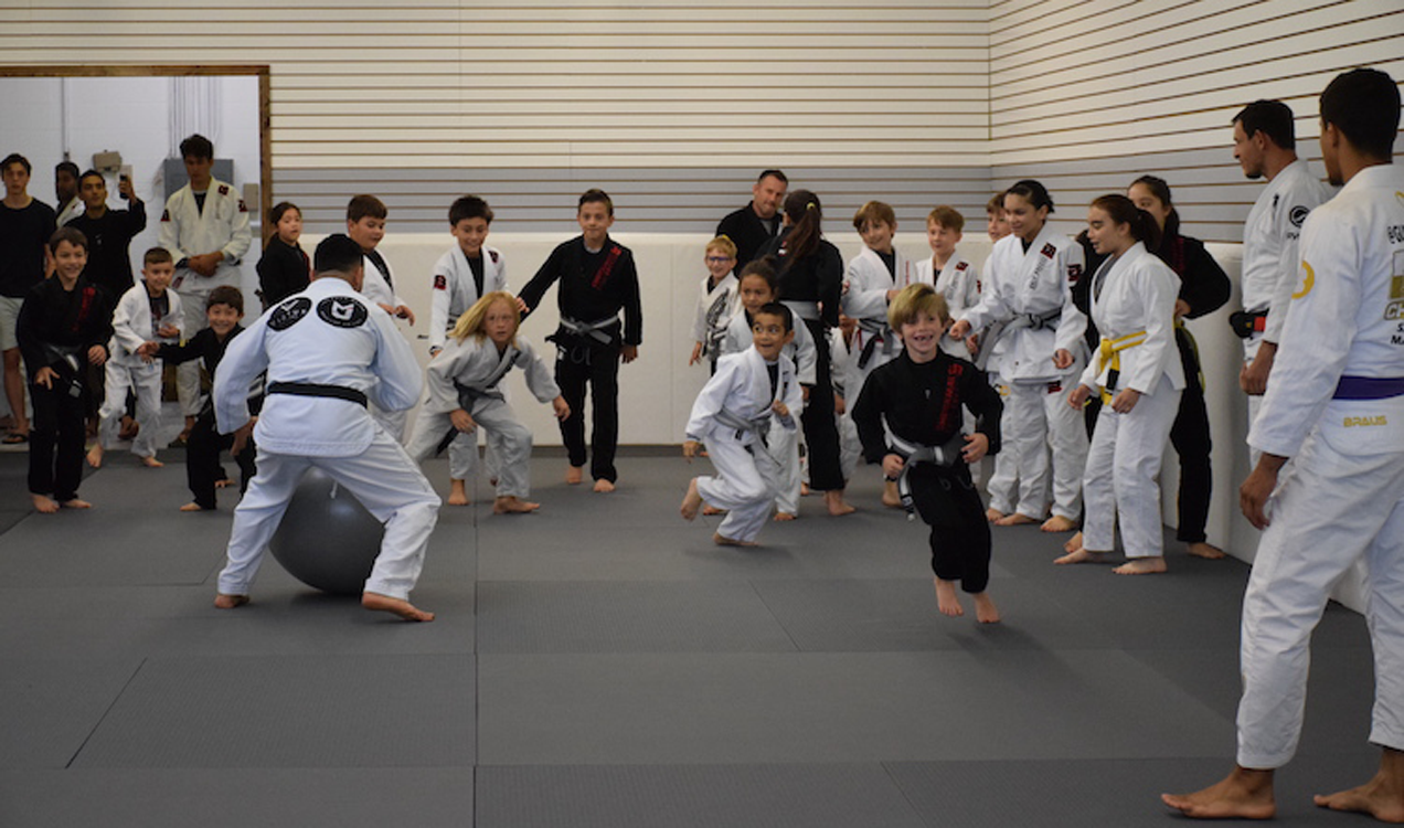 Kids Brazilian Jiu Jitsu class playing a fun training game on the mats with instructors supervising in a gym